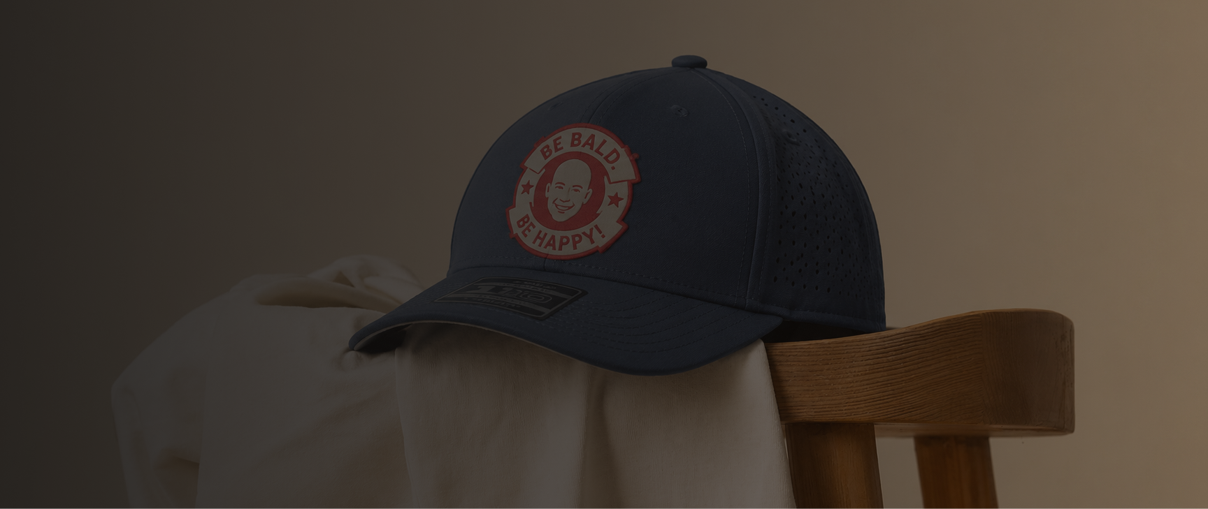 A dark navy blue baseball cap with a circular embroidered logo on the front, resting on a white draped cloth next to a wooden chair against a dark background.